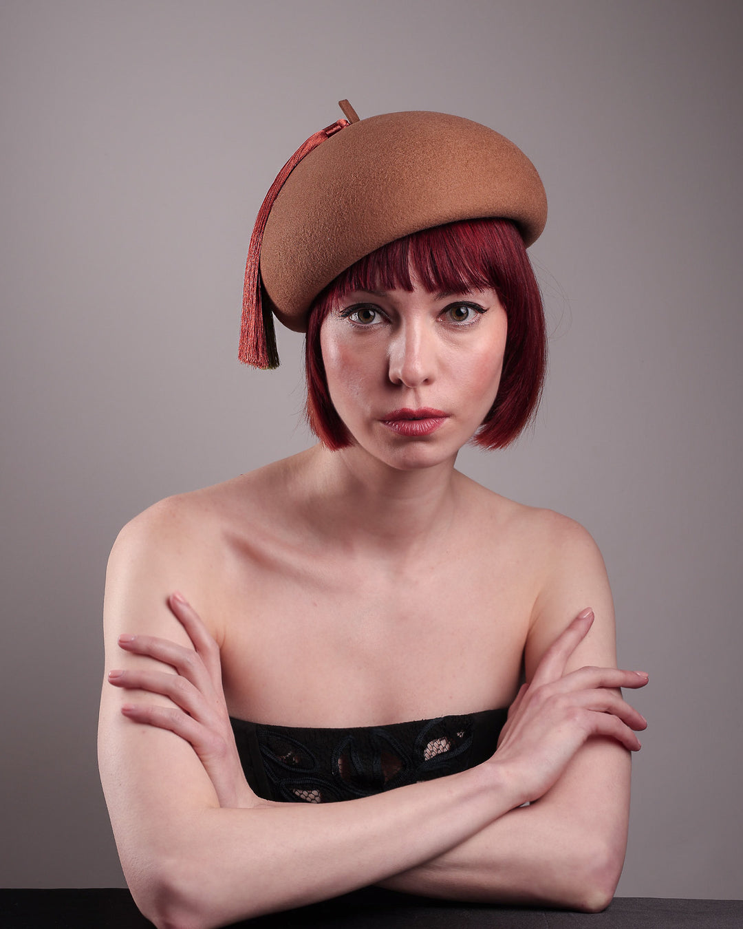 The model is showcasing the front view of the Faye Beret Hat, handcrafted in camel wool felt. It's clean, with a dome-shaped beret shape topped by an oversized dual-tone tassel in zesty chartreuse and burnt orange, creating a striking contrast against the warm neutral base. Designed by Merve Bayindir.