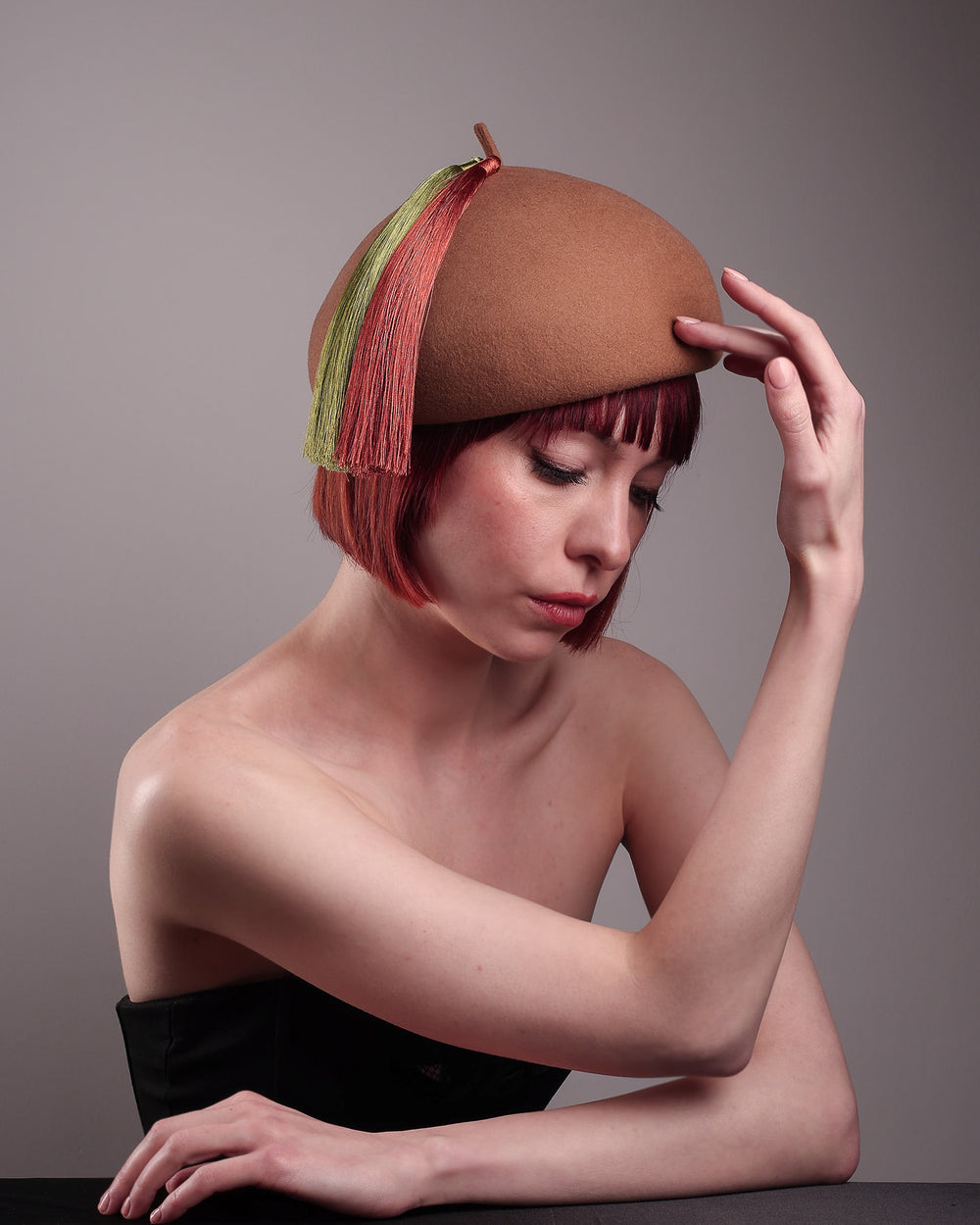 The model is showcasing the front right angle of the Faye Beret Hat, handcrafted in camel wool felt. It's clean, with a dome-shaped beret shape topped by an oversized dual-tone tassel in zesty chartreuse and burnt orange, creating a striking contrast against the warm neutral base. Designed by Merve Bayindir.