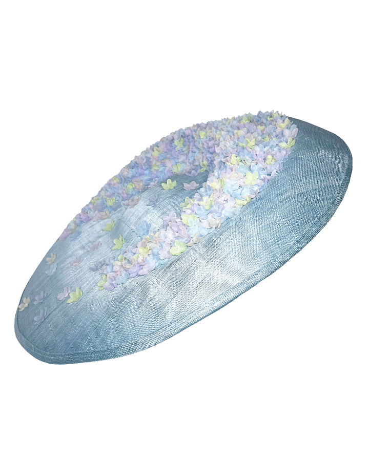 Powder blue sinamay base hat with a wide, gently sloping brim, adorned with clusters of soft pastel organza flowers in lilac, pale yellow, and blush tones that gather along the crown and trail lightly across the surface. Designed and made by Merve Bayindir Millinery.

