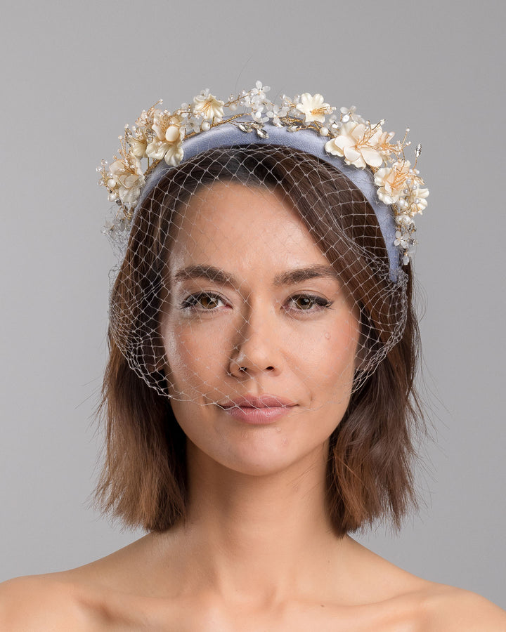 Model is showcasing the front close up of ice blue silk sateen headband decorated like crow with wired pearl colour flowers and light lilac veil. By Merve Bayindir Millinery.
