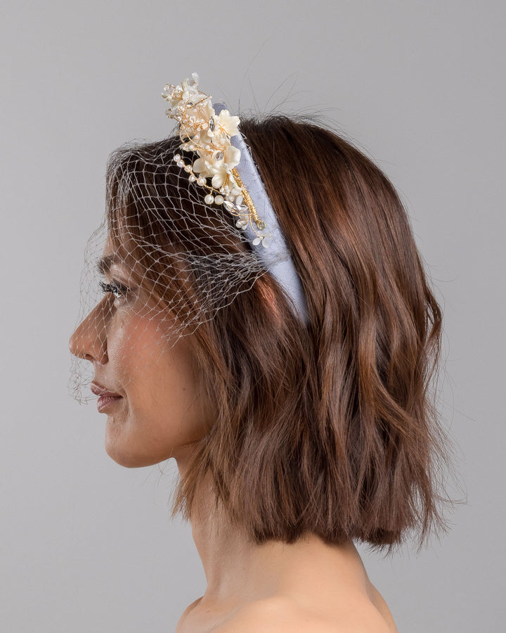 Model is showcasing the left side close up of ice blue silk sateen headband decorated like crow with wired pearl colour flowers and light lilac veil. By Merve Bayindir Millinery.