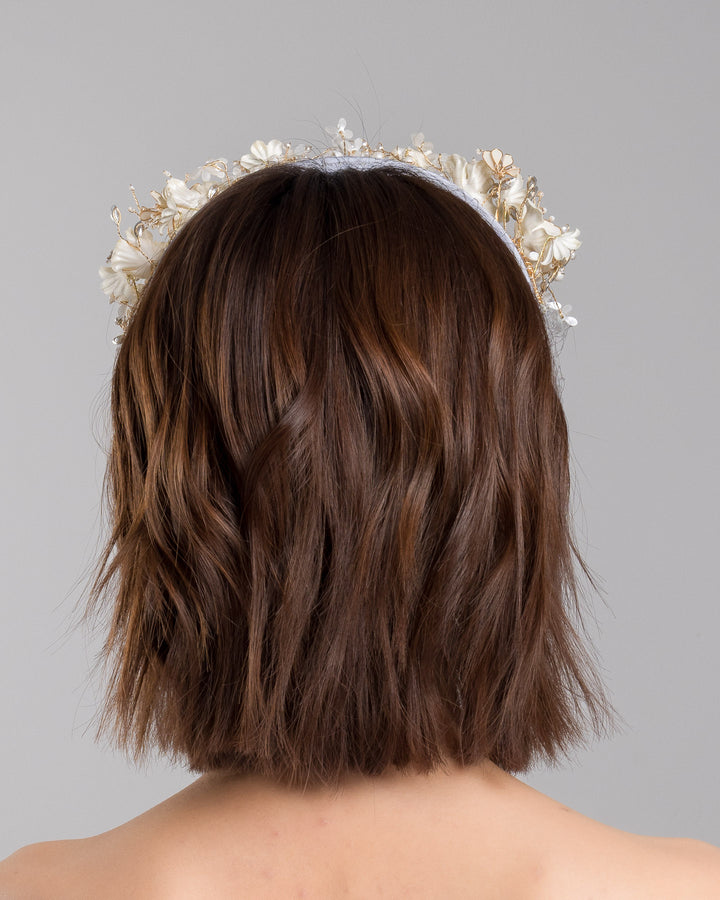 Model is showcasing the back side close up of ice blue silk sateen headband decorated like crow with wired pearl colour flowers and light lilac veil. By Merve Bayindir Millinery.