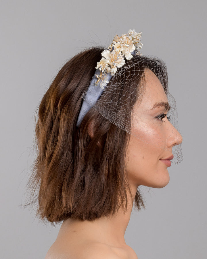 Model is showcasing the right side close up of ice blue silk sateen headband decorated like crow with wired pearl colour flowers and light lilac veil. By Merve Bayindir Millinery.