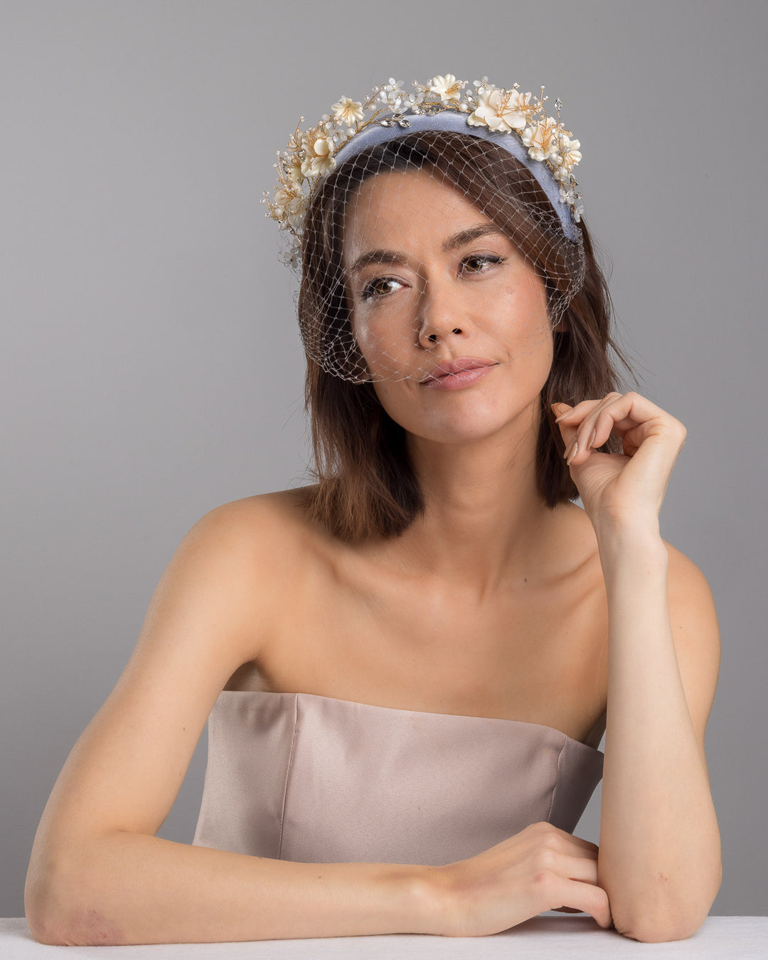 Model is posing wearing Fides ice blue silk sateen headband decorated like crow with wired pearl colour flowers and light lilac veil. By Merve Bayindir Millinery.