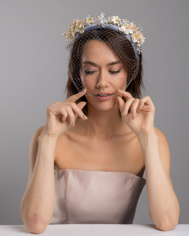 Model is posing wearing Fides ice blue silk sateen headband decorated like crow with wired pearl colour flowers and light lilac veil. By Merve Bayindir Millinery.