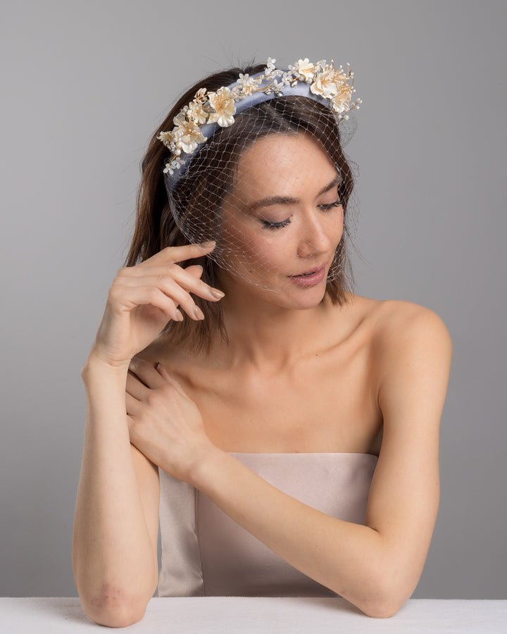 Model is posing wearing Fides ice blue silk sateen headband decorated like crow with wired pearl colour flowers and light lilac veil. By Merve Bayindir Millinery.