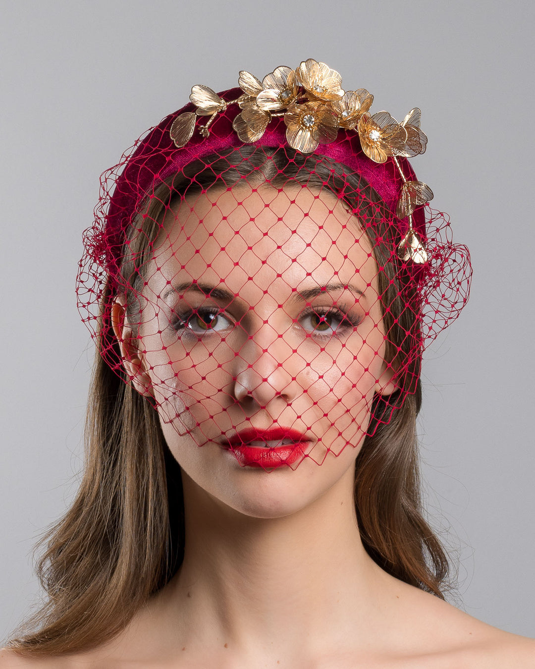 Model is showcasing the front of the wine coloured velvet headband declared with laser cut gold coloured wire flowers and finished with a red veil. Made by Merve bayindir millinery.