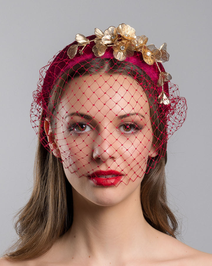 Model is showcasing the front of the wine coloured velvet headband declared with laser cut gold coloured wire flowers and finished with a red veil. Made by Merve bayindir millinery.