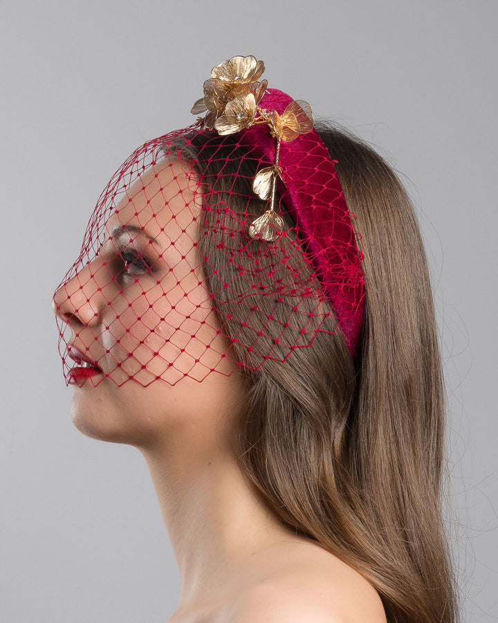 Model is showcasing the left side of the wine coloured velvet headband declared with laser cut gold coloured wire flowers and finished with a red veil. Made by Merve bayindir millinery.