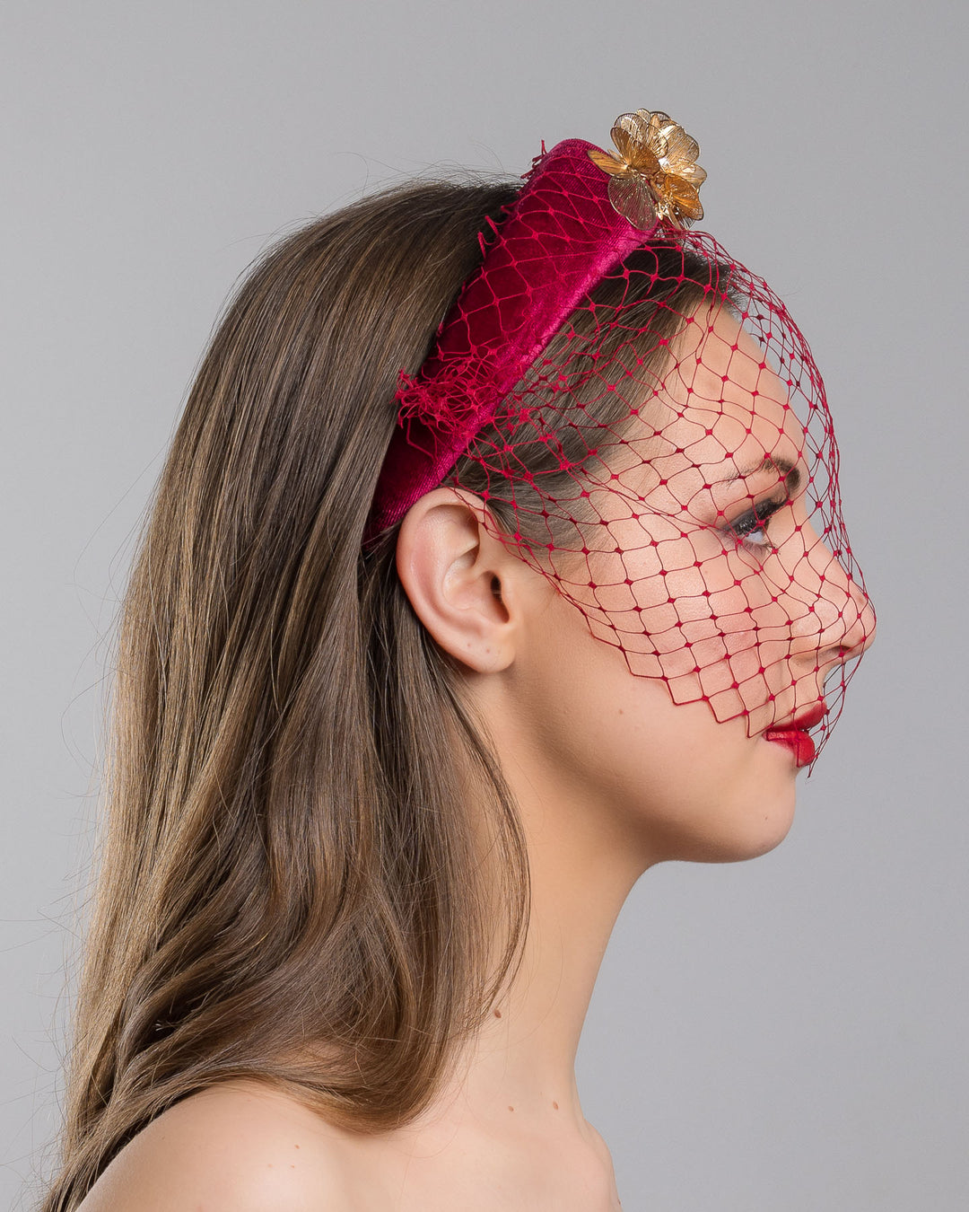 Model is showcasing the right side of the wine coloured velvet headband declared with laser cut gold coloured wire flowers and finished with a red veil. Made by Merve bayindir millinery.