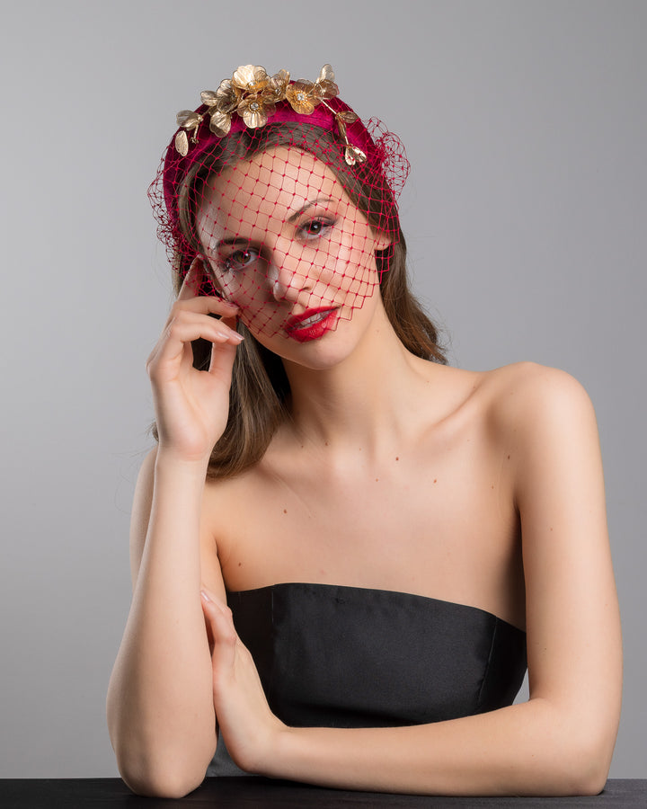 Model is posing wearing the wine coloured Fiducia velvet headband declared with laser cut gold coloured wire flowers and finished with a red veil. Made by Merve bayindir millinery.