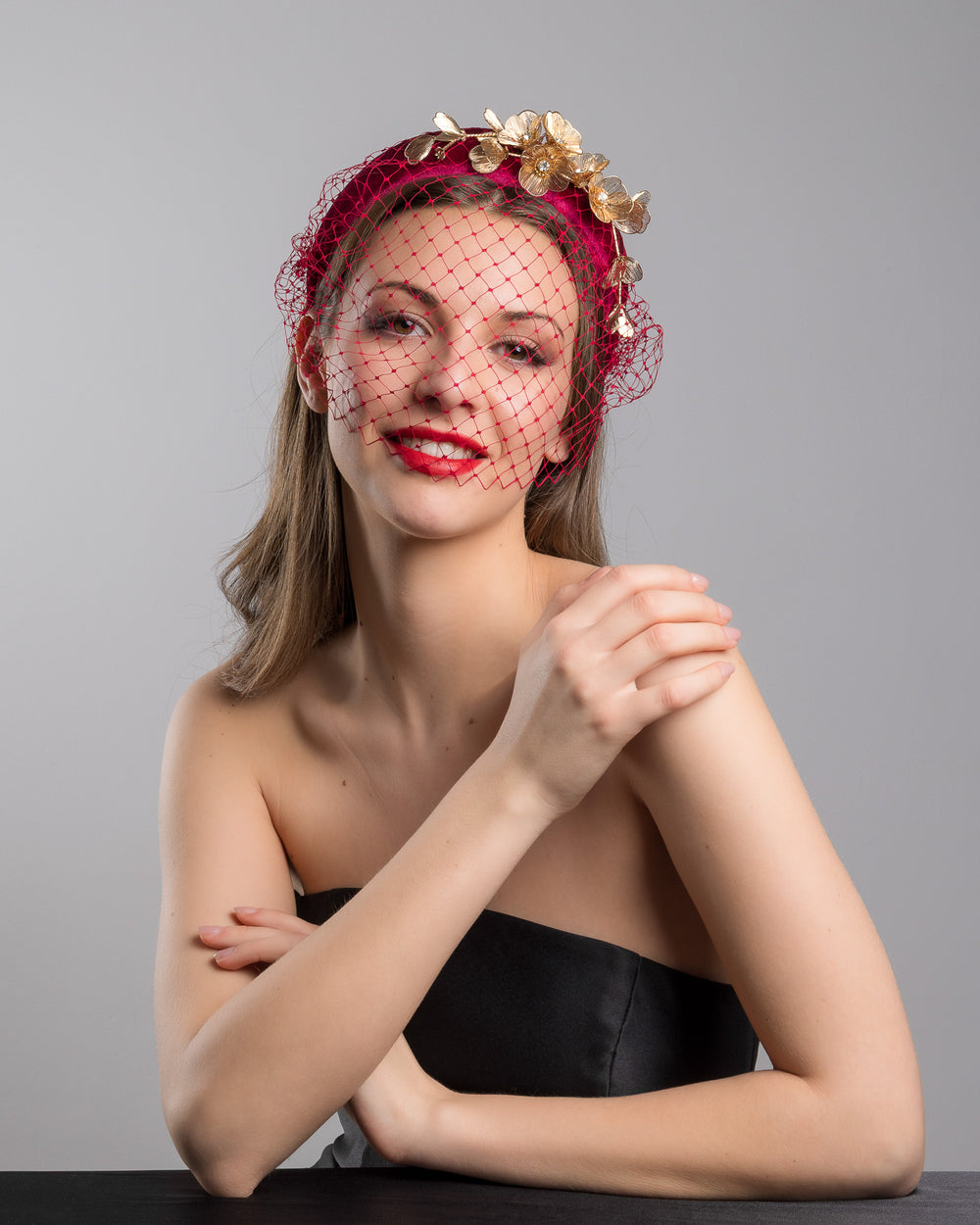 Model is posing wearing the wine coloured Fiducia velvet headband declared with laser cut gold coloured wire flowers and finished with a red veil. Made by Merve bayindir millinery.