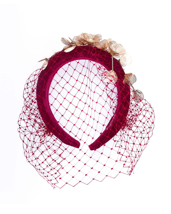 Still life picture of the wine coloured velvet headband declared with laser cut gold coloured wire flowers and finished with a red veil. Made by Merve bayindir millinery.