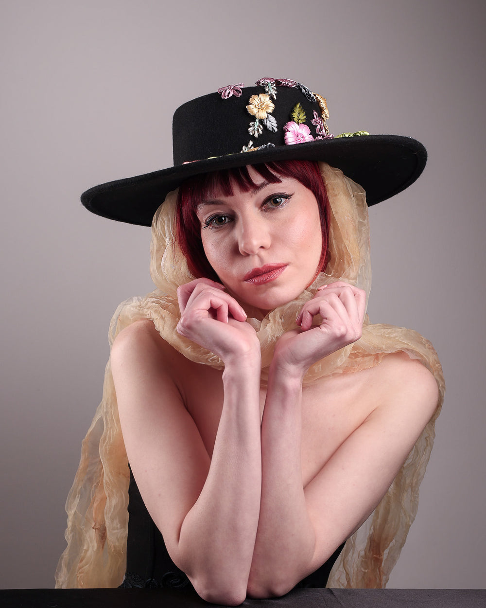 The female model is wearing a wool felt wide-brimmed boater hat showcasing the front view of the hat. The hat has multi-colour satin embroidery flower details on the right side of the hat, as well as an organza long scarf that falls from both sides. Designed by Merve Bayindir.