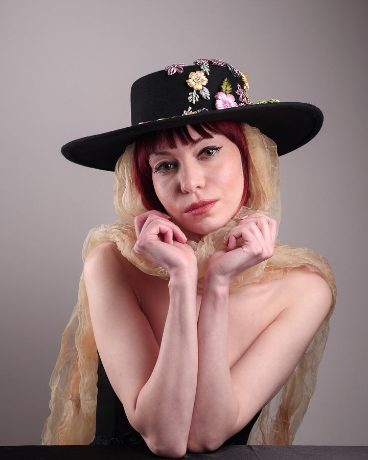 The female model is wearing a wool felt wide-brimmed boater hat showcasing the front view of the hat. The hat has multi-colour satin embroidery flower details on the right side of the hat, as well as an organza long scarf that falls from both sides. Designed by Merve Bayindir.