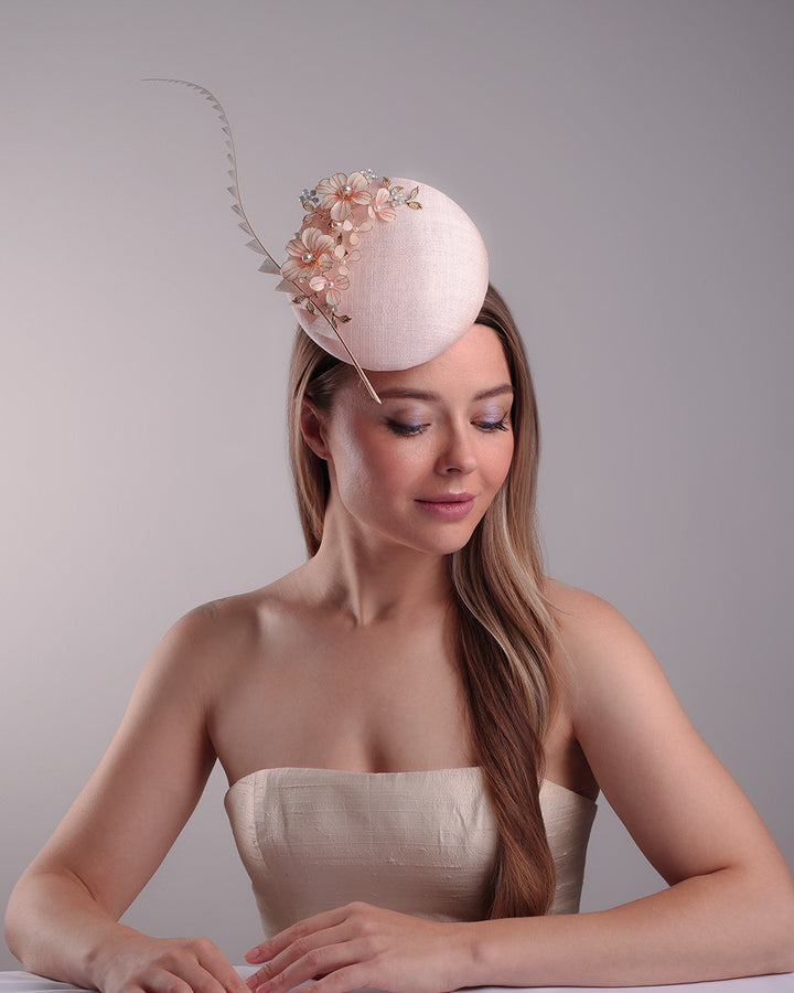 The model presents the double sided high button shaped pink sinamay fascinator, placed on the right front side of the head, decorated with wired resin flowers and crystal bouquet and a single trimmed rooster feather on the side. The piece is fitted with a custom metal headband and elastic.