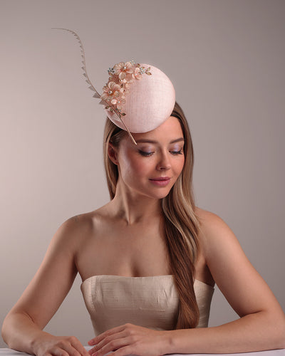 The model presents the double sided high button shaped pink sinamay fascinator, placed on the right front side of the head, decorated with wired resin flowers and crystal bouquet and a single trimmed rooster feather on the side. The piece is fitted with a custom metal headband and elastic.