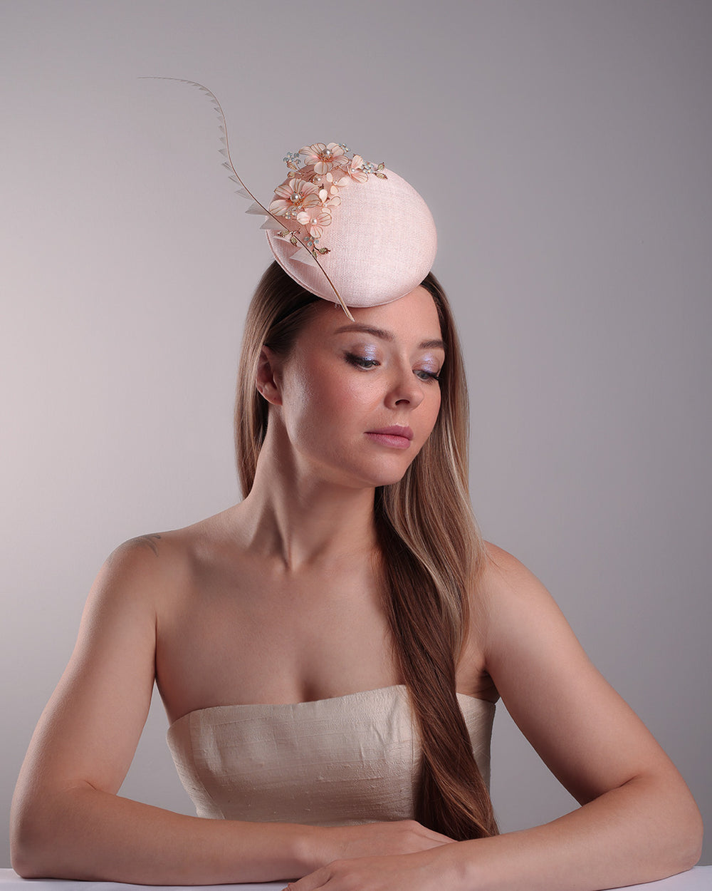 The model presents the right front angle of the high button shaped pink sinamay fascinator, placed on the right front side of the head, decorated with wired resin flowers and crystal bouquet and a single trimmed rooster feather on the side. The piece is fitted with a custom metal headband and elastic.