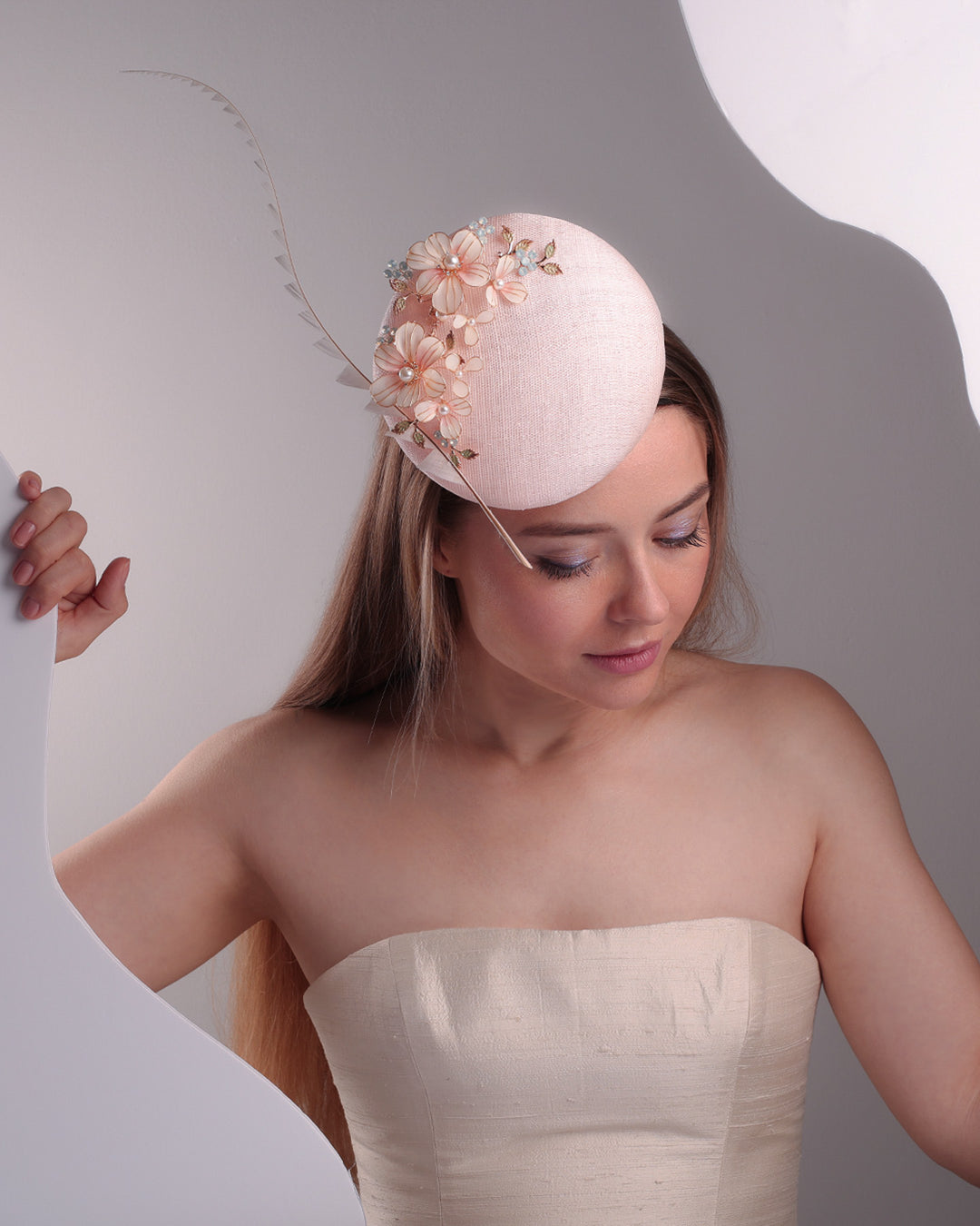 The model presents the double sided high button shaped pink sinamay fascinator from the above, placed on the right front side of the head, decorated with wired resin flowers and crystal bouquet and a single trimmed rooster feather on the side. The piece is fitted with a custom metal headband and elast