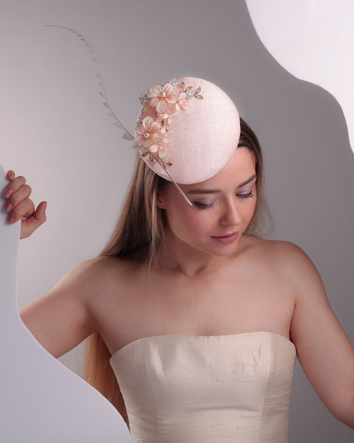 The model presents the double sided high button shaped pink sinamay fascinator from the above, placed on the right front side of the head, decorated with wired resin flowers and crystal bouquet and a single trimmed rooster feather on the side. The piece is fitted with a custom metal headband and elast