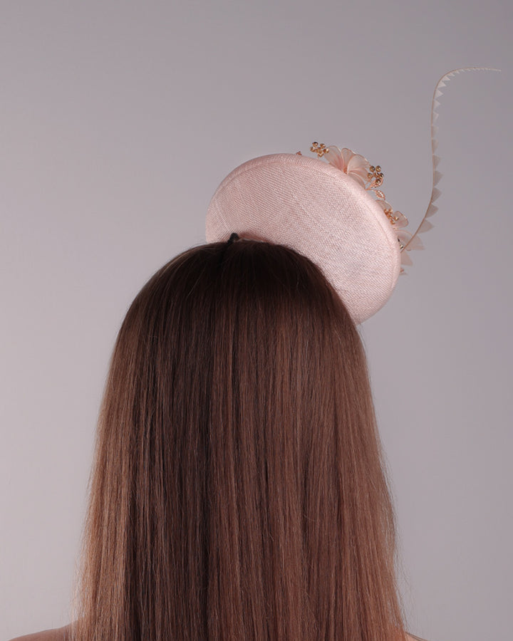 The model presents the back view of the double sided high button shaped pink sinamay fascinator, placed on the right front side of the head, decorated with wired resin flowers and crystal bouquet and a single trimmed rooster feather on the side. The piece is fitted with a custom metal headband and elastic.