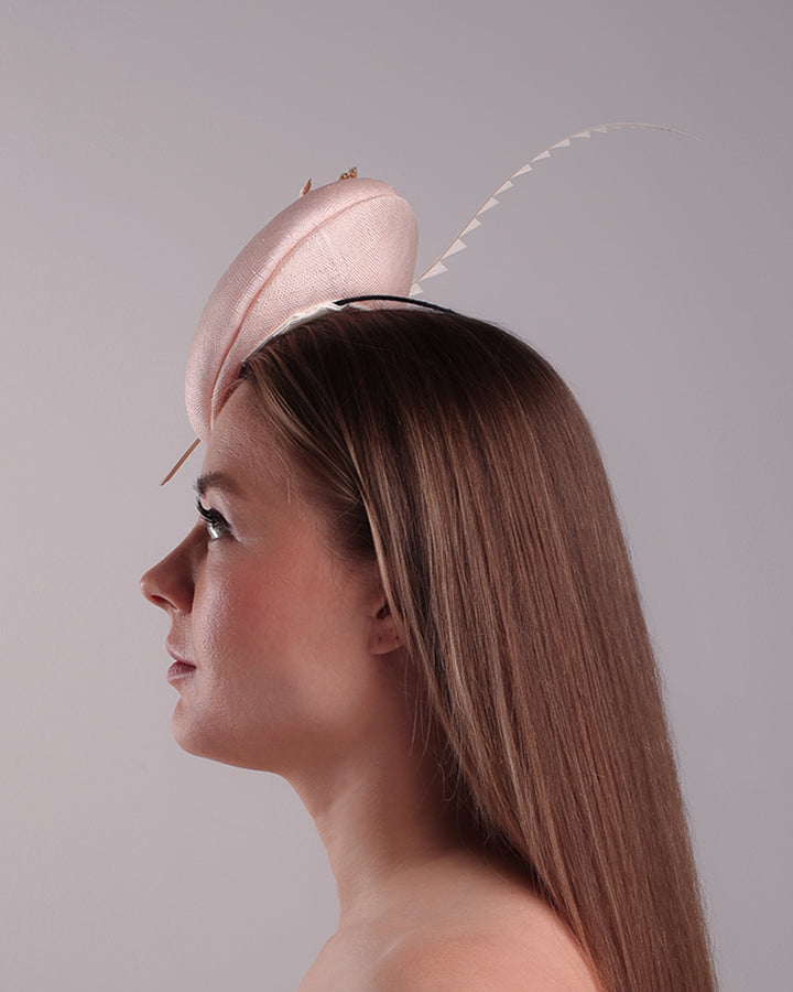 The model presents the left side of  the double sided high button shaped pink sinamay fascinator, placed on the right front side of the head, decorated with wired resin flowers and crystal bouquet and a single trimmed rooster feather on the side. The piece is fitted with a custom metal headband and elastic.