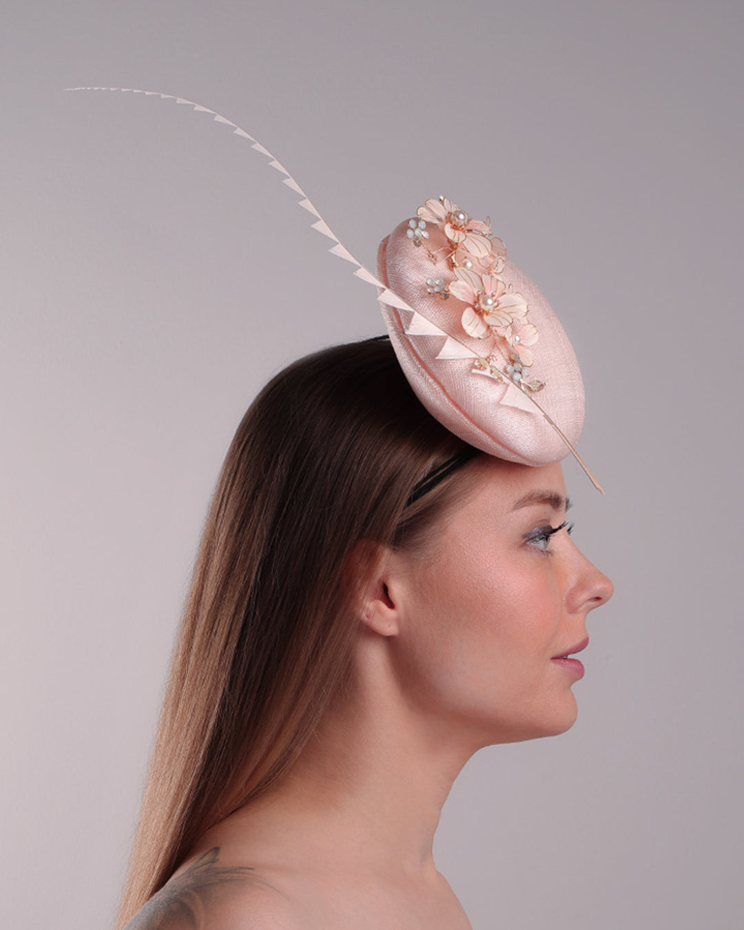 The model presents the right side of the double sided high button shaped pink sinamay fascinator, placed on the right front side of the head, decorated with wired resin flowers and crystal bouquet and a single trimmed rooster feather on the side. The piece is fitted with a custom metal headband and elastic.