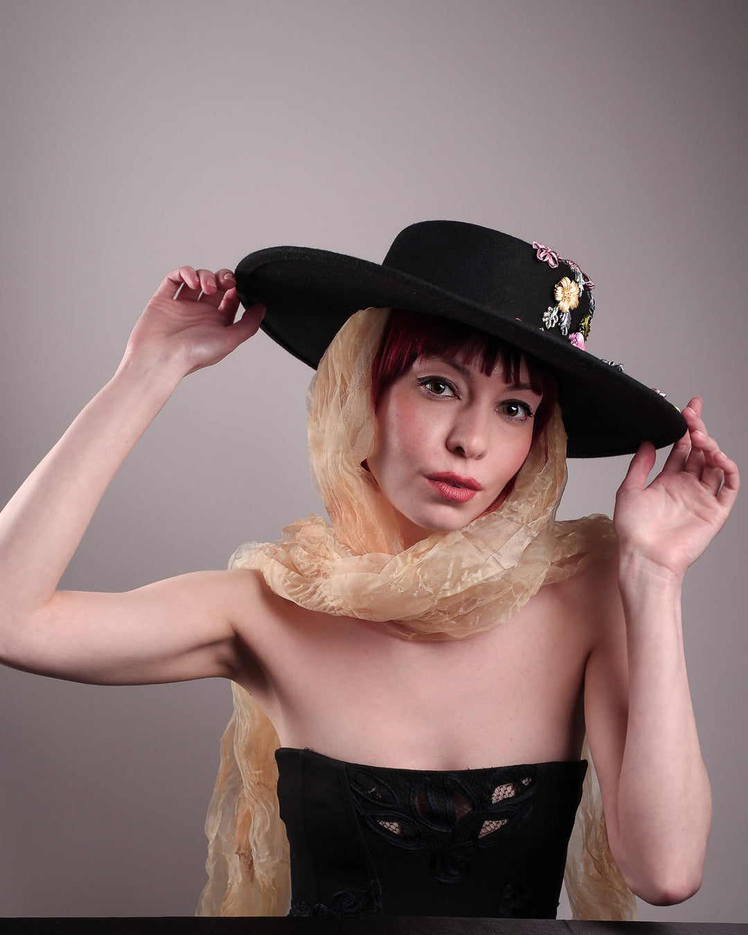 The female model is showcasing a wool felt wide-brimmed boater hat from the front below angle. The hat has multi-colour satin embroidery flower details on the right side of the hat, as well as an organza long scarf that falls from both sides. Designed by Merve Bayindir.