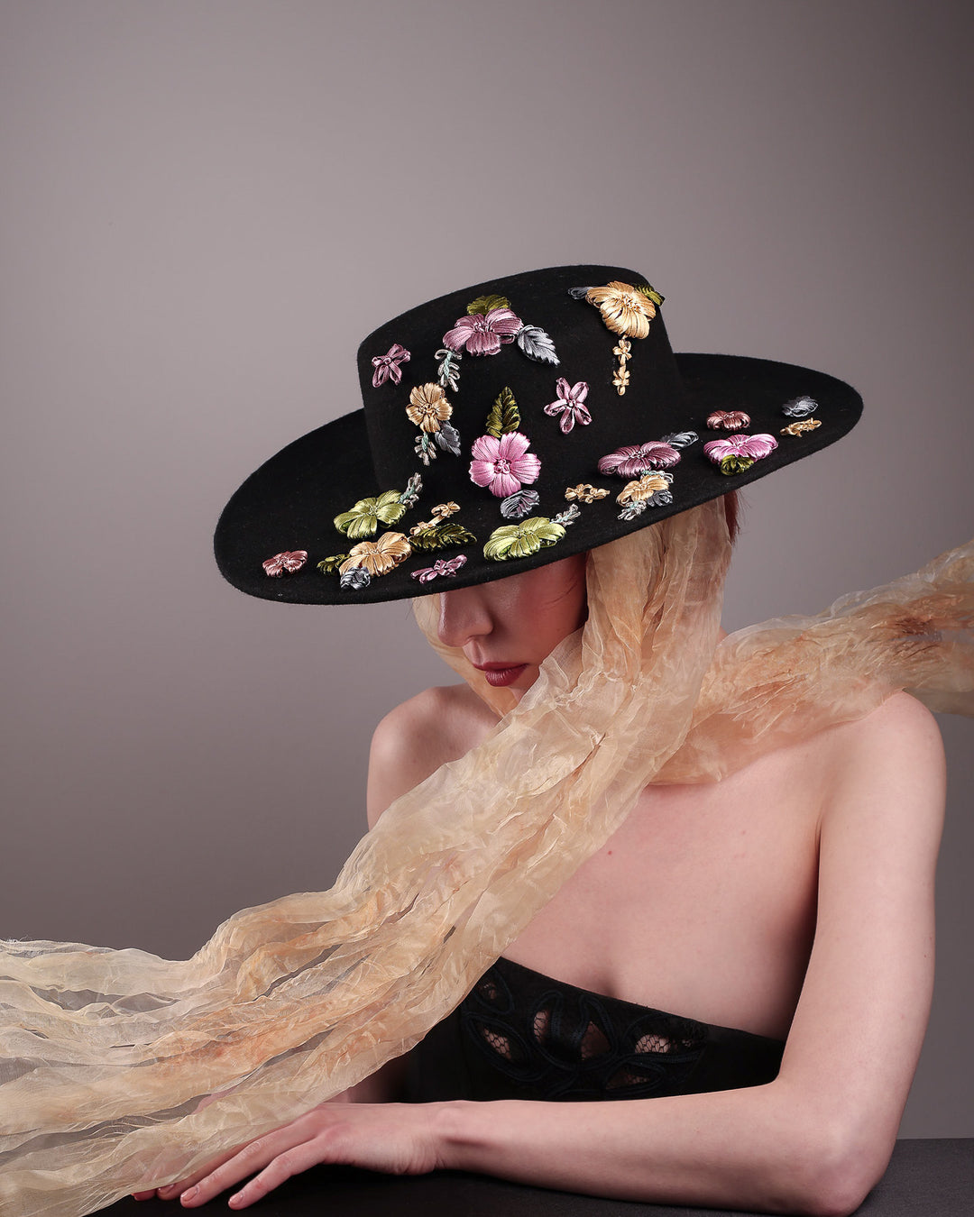 The female model is wearing a wool felt wide-brimmed boater hat. The right angled picture showcasing the front the right side of the hat which has multi-colour satin embroidery flower details, as well as the length of an organza long scarf that falls from both sides. Designed by Merve Bayindir.