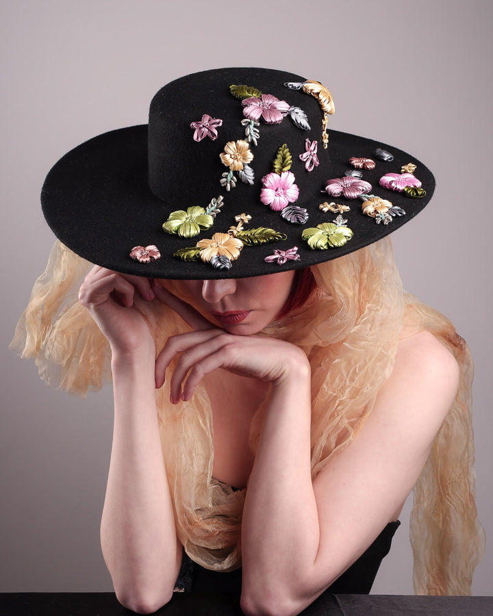 The female wearing a wool felt wide-brimmed boater hat, the above angled picture showcases the detailed view of the  multi-colour satin embroidery flower details on the right side of the hat, as well as an organza long scarf that falls from both sides. Designed by Merve Bayindir.