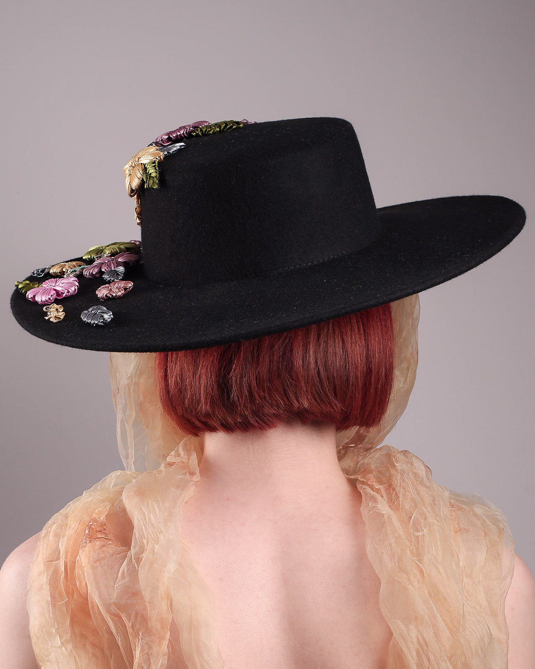 The female model is showcasing the back of the wool felt wide-brimmed boater hat. The right side of the hat which has multi-colour satin embroidery flower details, as well as the length of an organza long scarf that falls from both sides. Designed by Merve Bayindir.