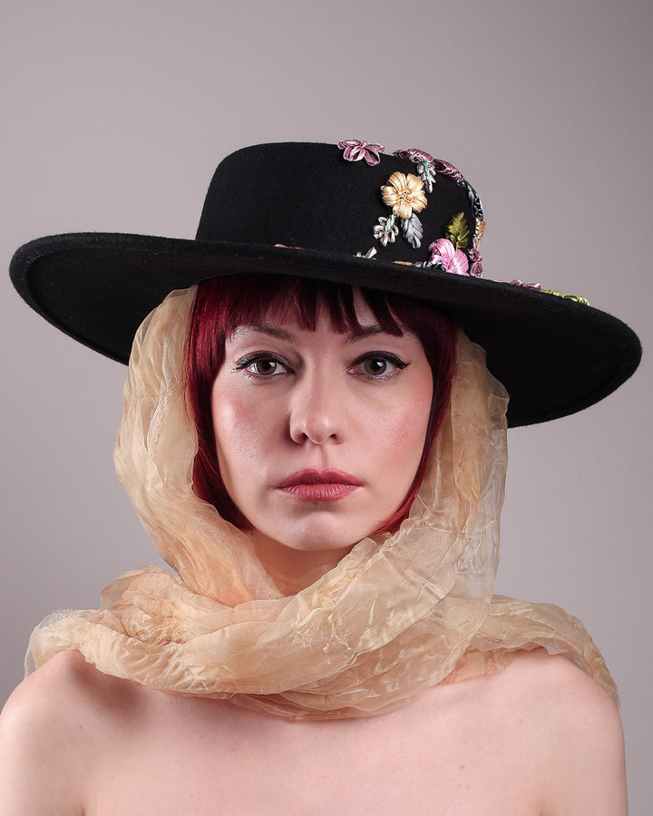 The female model is showcasing the front close up view  a wool felt wide-brimmed boater hat. The right side of the hat which has multi-colour satin embroidery flower details, as well as the length of an organza long scarf that falls from both sides. Designed by Merve Bayindir.