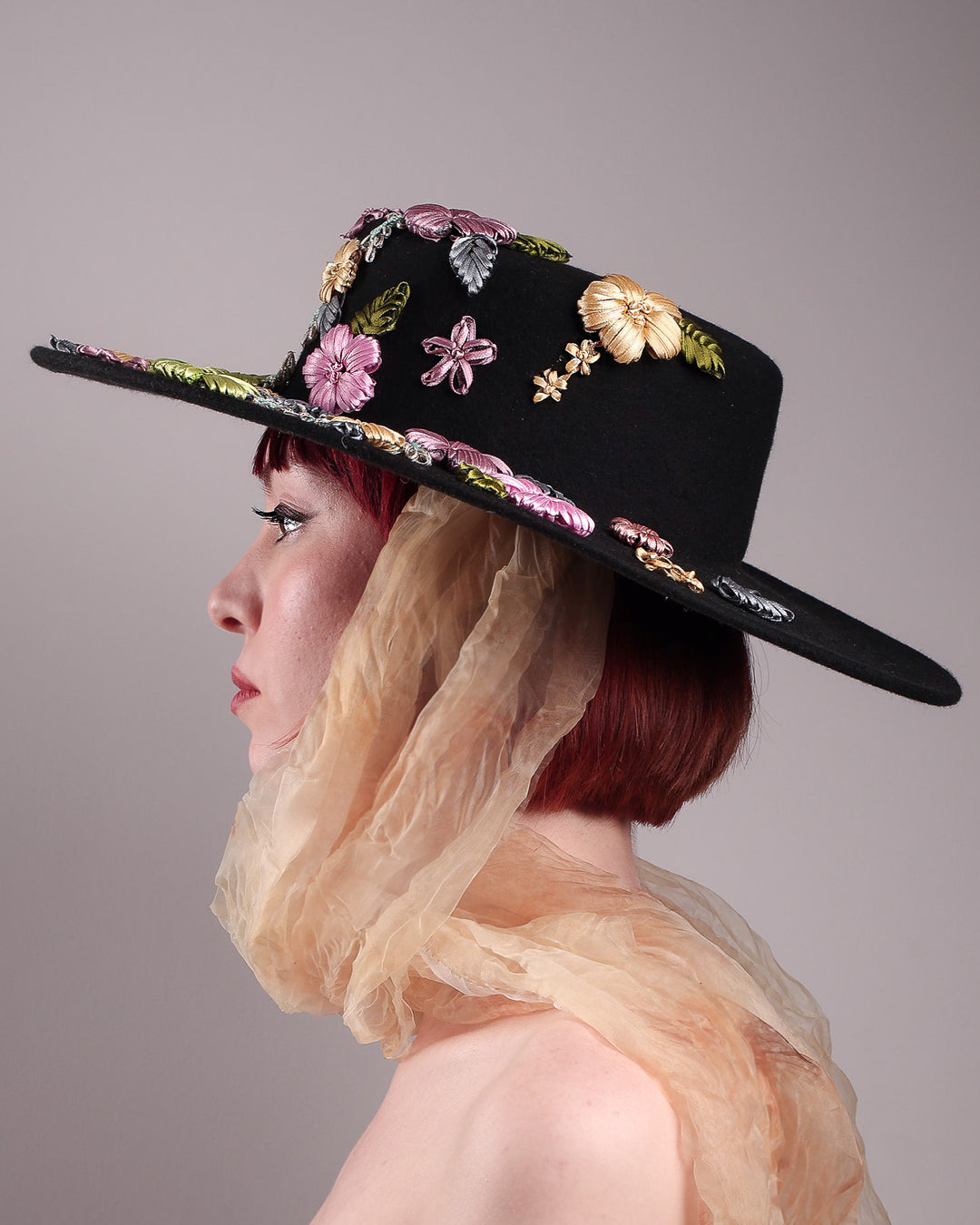 The female model is showcasing the right side close up view  a wool felt wide-brimmed boater hat. The right side of the hat which has multi-colour satin embroidery flower details, as well as the length of an organza long scarf that falls from both sides. Designed by Merve Bayindir.