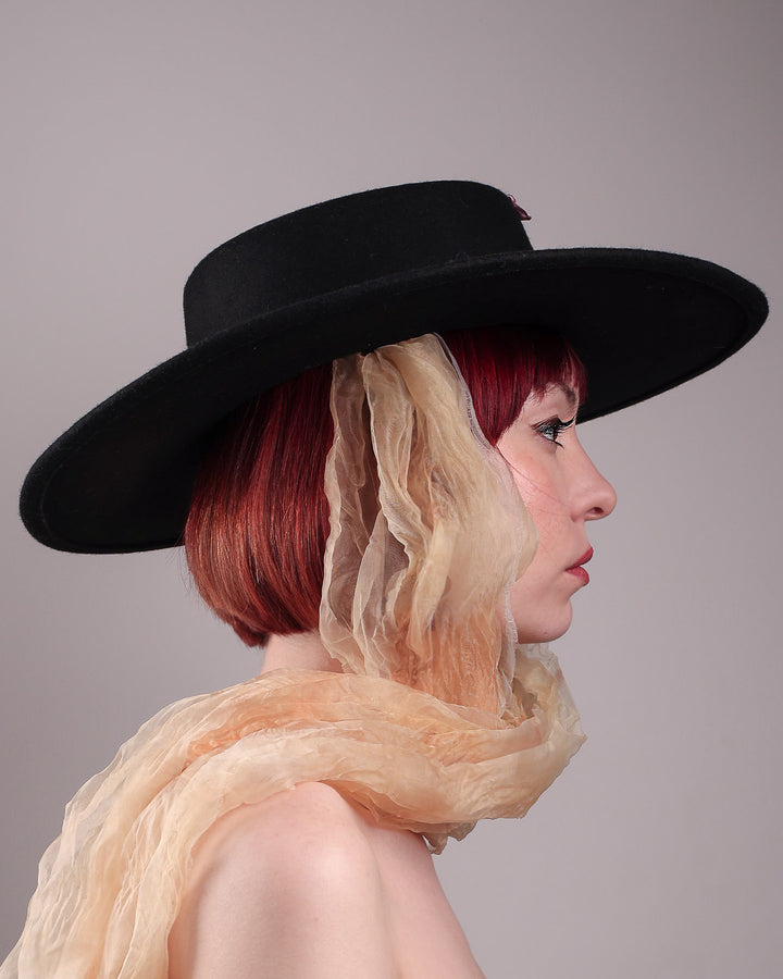 The female model is showcasing the left side close up view  a wool felt wide-brimmed boater hat. The right side of the hat which has multi-colour satin embroidery flower details, as well as the length of an organza long scarf that falls from both sides. Designed by Merve Bayindir.