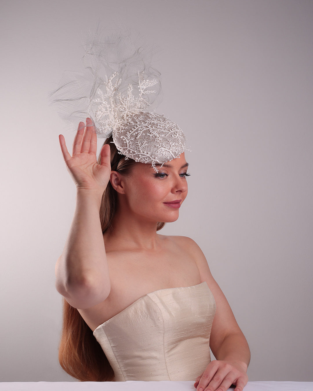 Model is showcasing the right front part of a grey button shaped fascinator, fully covered with white branch look designed 3D lace with movement. Embellished with a grey crinoline cloud detaling a wired lace pieaces looking like branches. Fitted with a wire heaband and elastic.