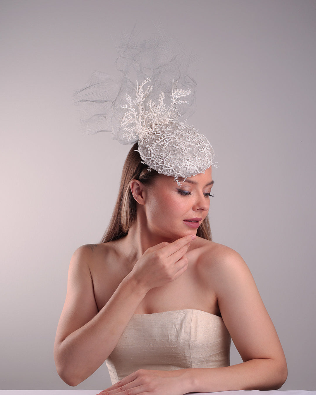 Model is showcasing the right top front part of a grey button shaped fascinator, fully covered with white branch look designed 3D lace with movement. Embellished with a grey crinoline cloud detaling a wired lace pieaces looking like branches. Fitted with a wire heaband and elastic.