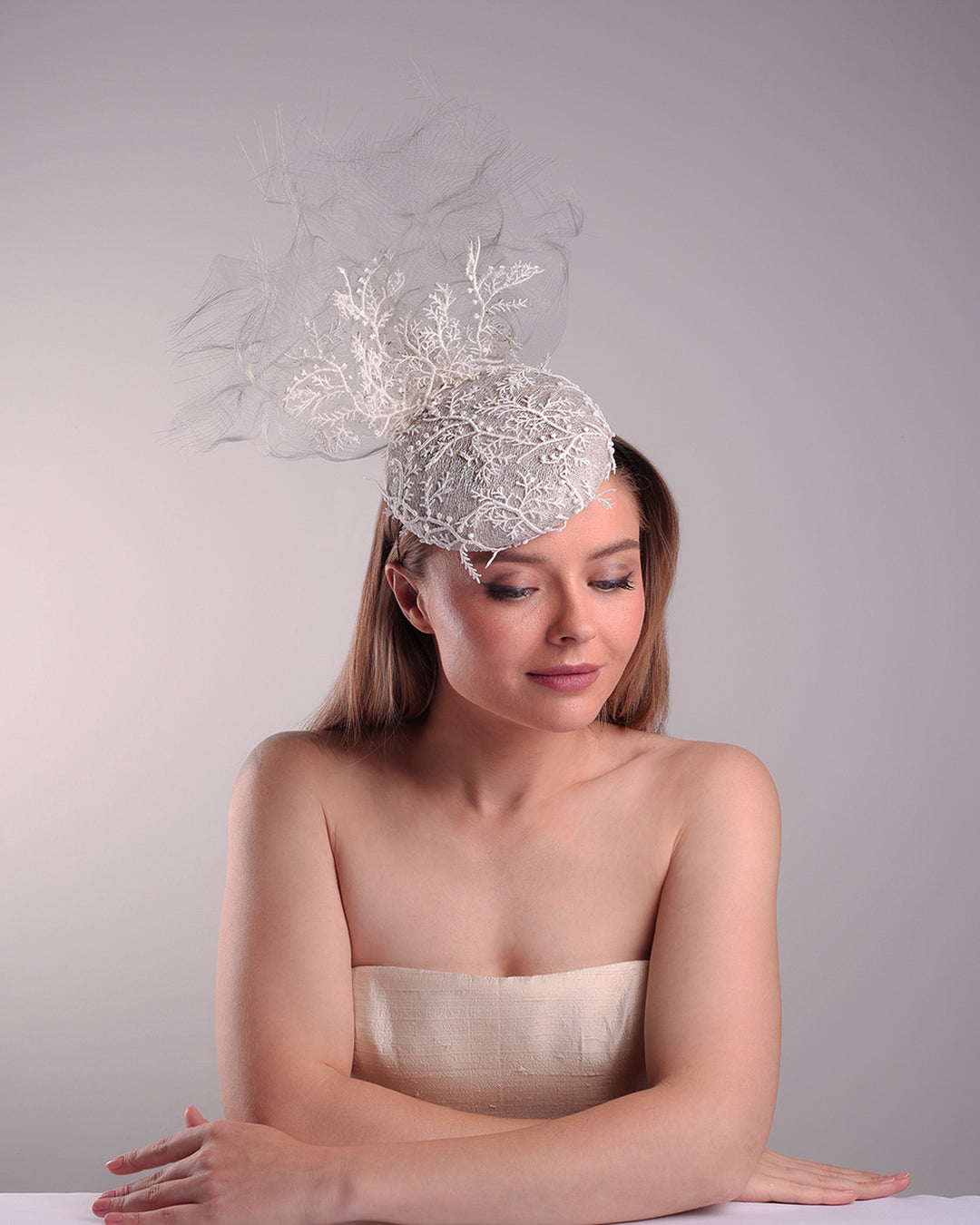 Model is showcasing the top front part of a grey button shaped fascinator, fully covered with white branch look designed 3D lace with movement. Embellished with a grey crinoline cloud detaling a wired lace pieaces looking like branches. Fitted with a wire heaband and elastic.