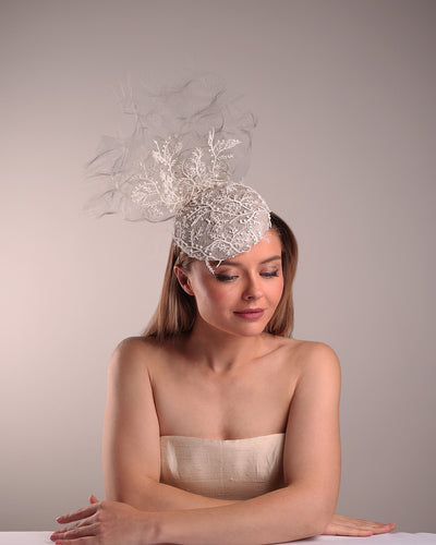 Model is showcasing the top front part of a grey button shaped fascinator, fully covered with white branch look designed 3D lace with movement. Embellished with a grey crinoline cloud detaling a wired lace pieaces looking like branches. Fitted with a wire heaband and elastic.