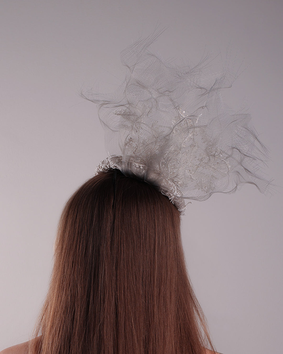 Model is showcasing the back of a grey button shaped fascinator, fully covered with white branc look designed 3D lace with movement. Embellished with a grey crinoline cloud detaling a wired lace pieaces looking like branches. Fitted with a wire heaband and elastic.