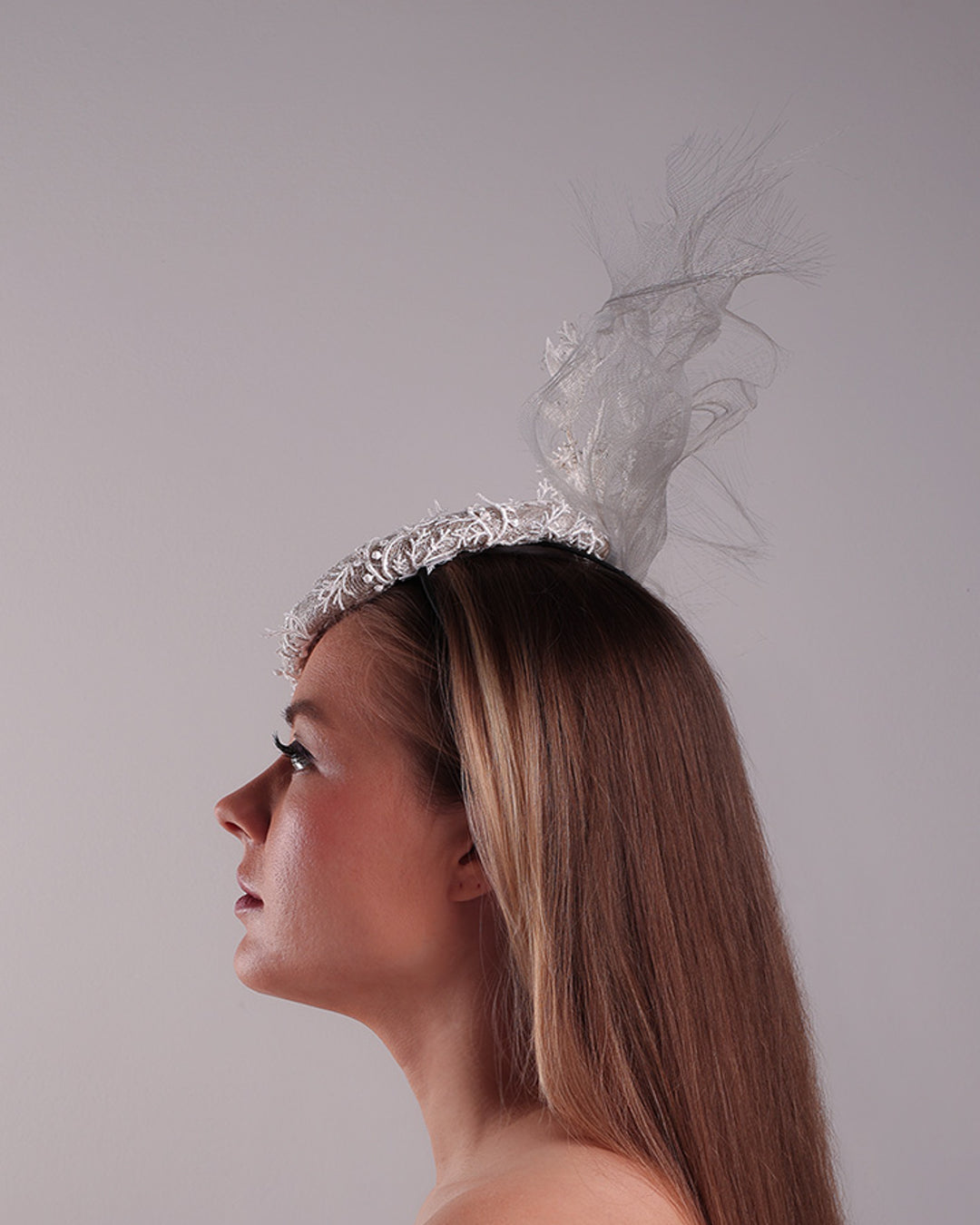 Model is showcasing the left side of a grey button shaped fascinator, fully covered with white branc look designed 3D lace with movement. Embellished with a grey crinoline cloud detaling a wired lace pieaces looking like branches. Fitted with a wire heaband and elastic.