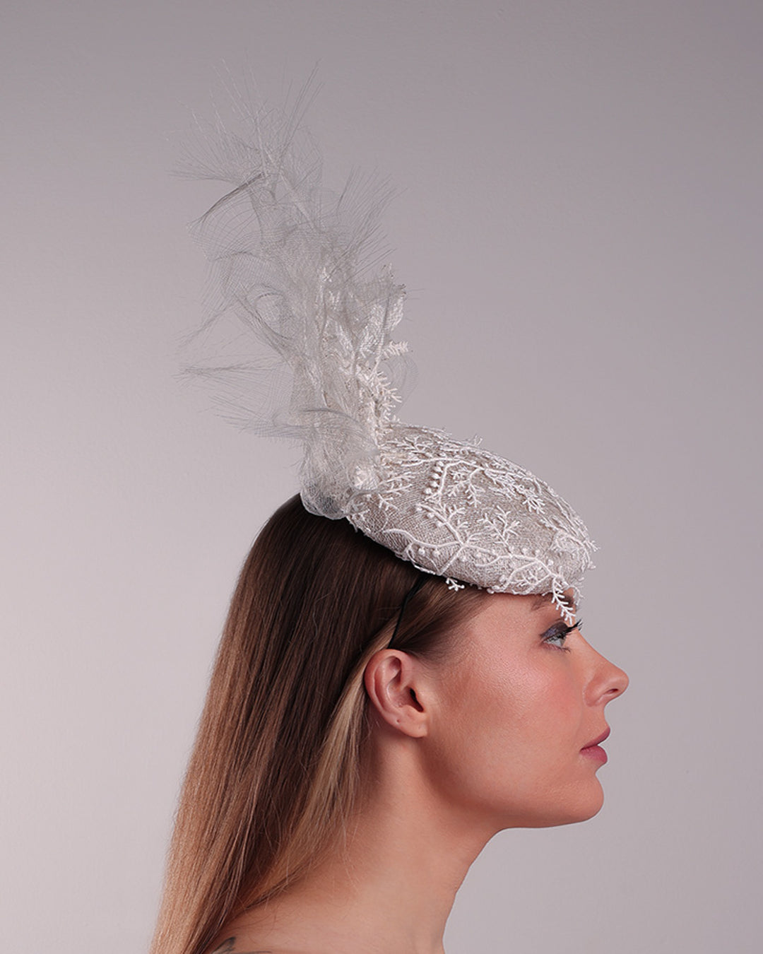 Model is showcasing the back of a grey button shaped fascinator, fully covered with white branc look designed 3D lace with movement. Embellished with a grey crinoline cloud detaling a wired lace pieaces looking like branches. Fitted with a wire heaband and elastic.