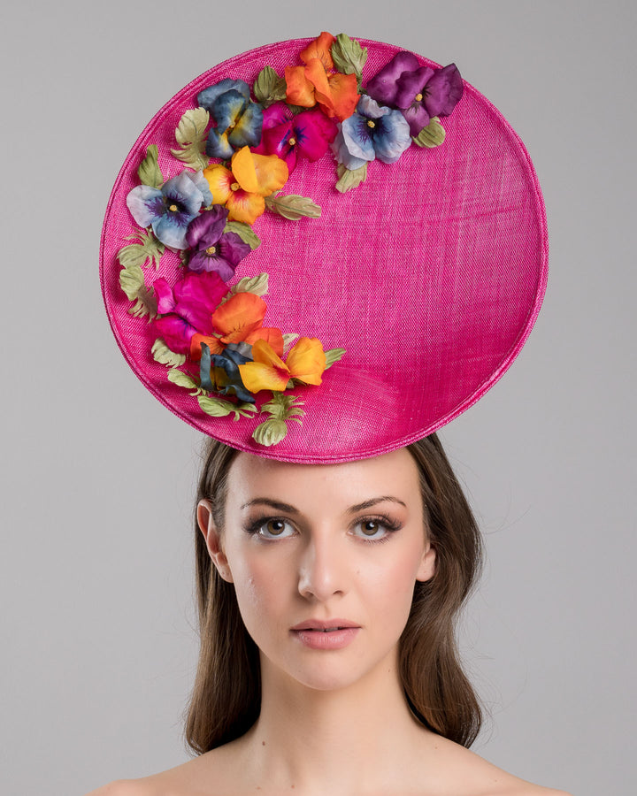 The model is showcasing the front side close up of the large size oval shaped hot pink fascinator decorated with custom made silk organza multi-coloured violets. Designed and made by Merve Bayindir Millinery.
