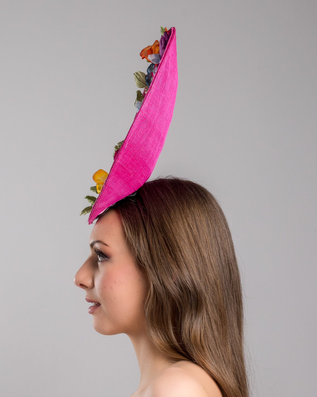 The model is showcasing the left side close up of the large size oval shaped hot pink fascinator decorated with custom made silk organza multi-coloured violets. Designed and made by Merve Bayindir Millinery.

