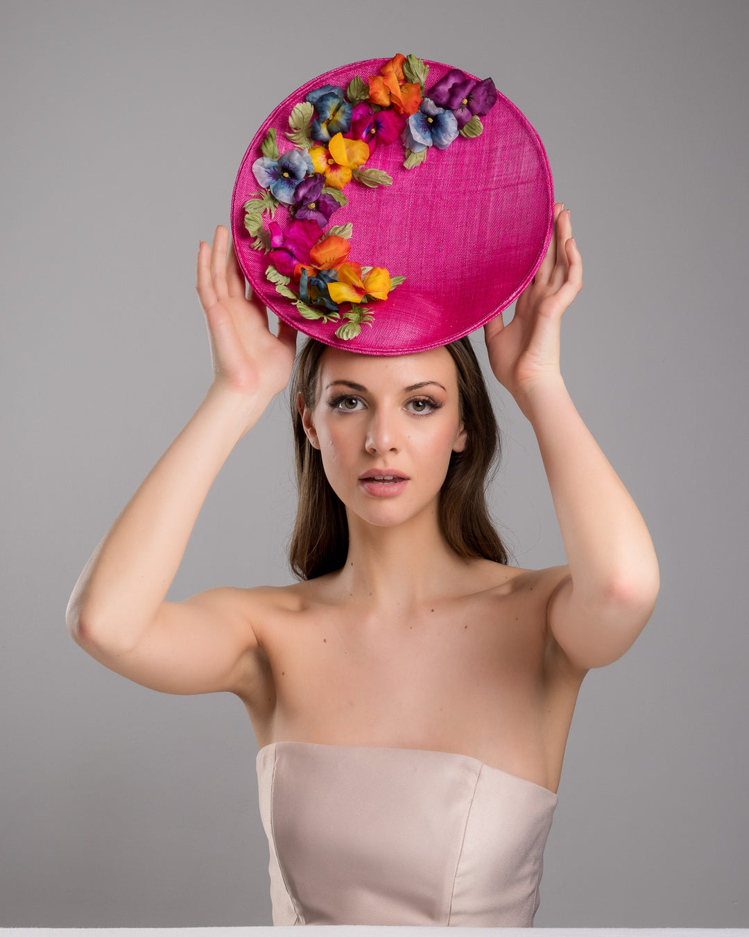 The model is posing, wearing GOZO,  large size oval shaped hot pink fascinator decorated with custom made silk organza multi-coloured violets. Designed and made by Merve Bayindir Millinery.
