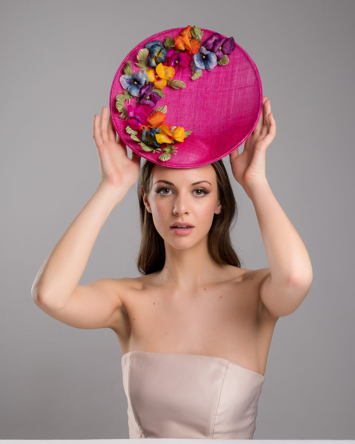 The model is posing, wearing GOZO,  large size oval shaped hot pink fascinator decorated with custom made silk organza multi-coloured violets. Designed and made by Merve Bayindir Millinery.
