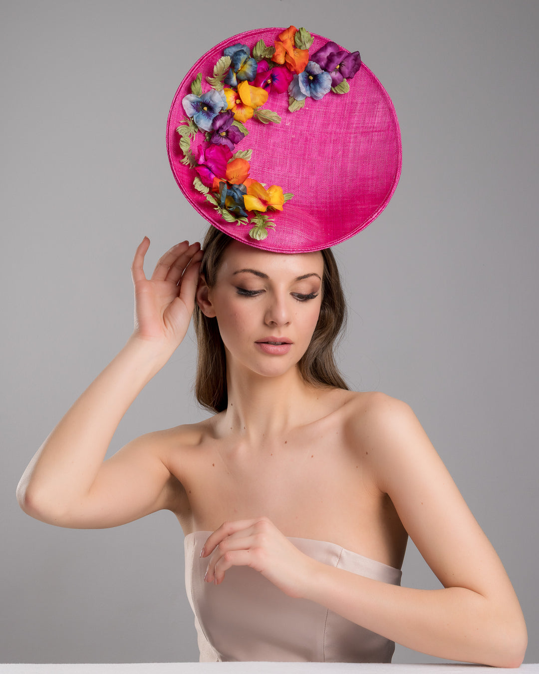 The model is posing, wearing GOZO,  large size oval shaped hot pink fascinator decorated with custom made silk organza multi-coloured violets. Designed and made by Merve Bayindir Millinery.
