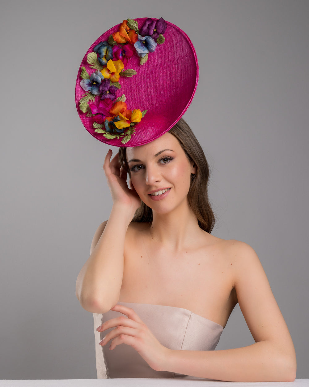 The model is posing, wearing GOZO,  large size oval shaped hot pink fascinator decorated with custom made silk organza multi-coloured violets. Designed and made by Merve Bayindir Millinery.
