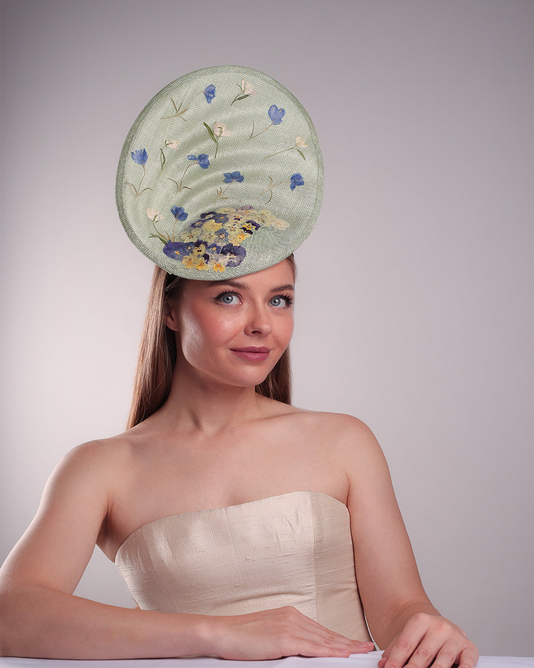 Model is showcasing the front of the light green tall fascinator with curves on the tall side like a sea shell, decorated with multi-colour pressed natural flowers. Fitted to the head with wire headband and elastic.