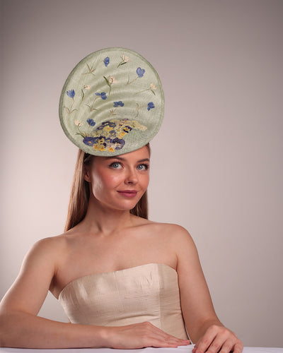 Model is showcasing the front of the light green tall fascinator with curves on the tall side like a sea shell, decorated with multi-colour pressed natural flowers. Fitted to the head with wire headband and elastic.