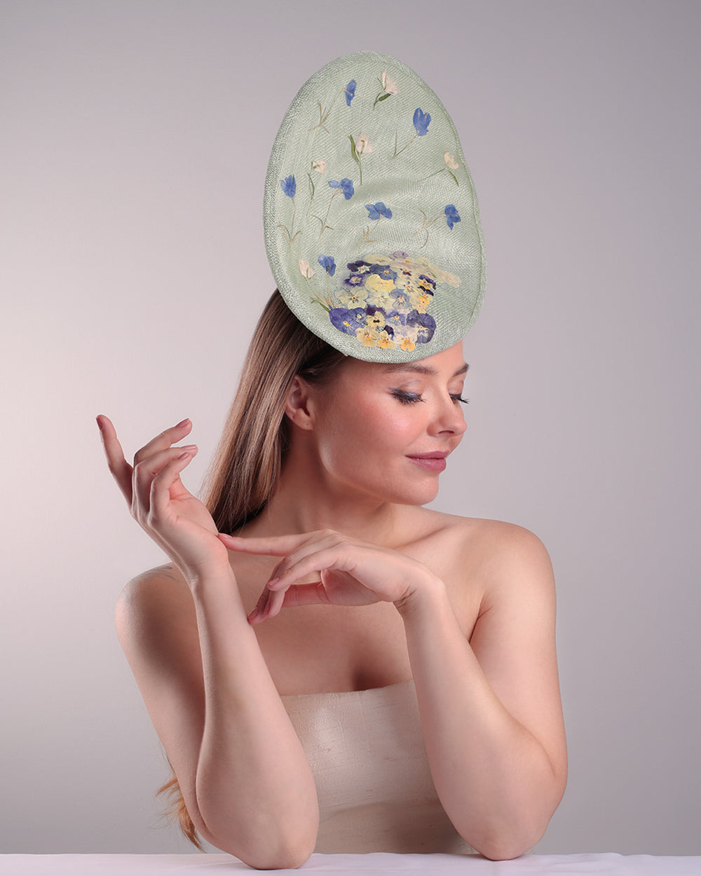 Model is showcasing the front right side of the light green tall fascinator with curves on the tall side like a sea shell, decorated with multi-colour pressed natural flowers. Fitted to the head with wire headband and elastic.